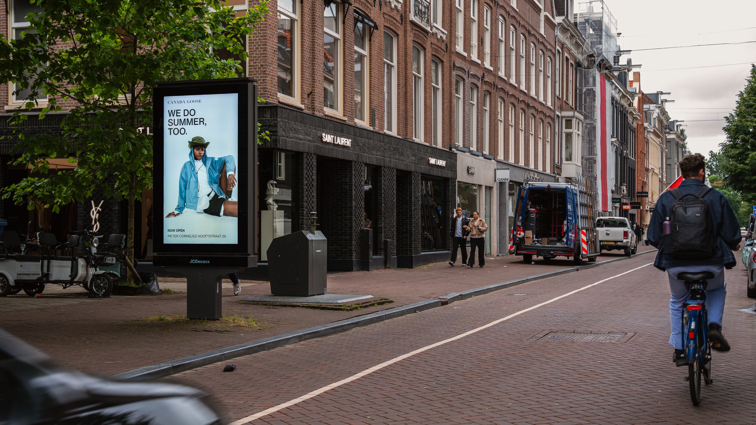 Stadsstraat met mensen, fietsen en winkels, waaronder Saint Laurent winkel, met een digitale reclamezuil naast een boom en een fietsende persoon in de voorgrond.