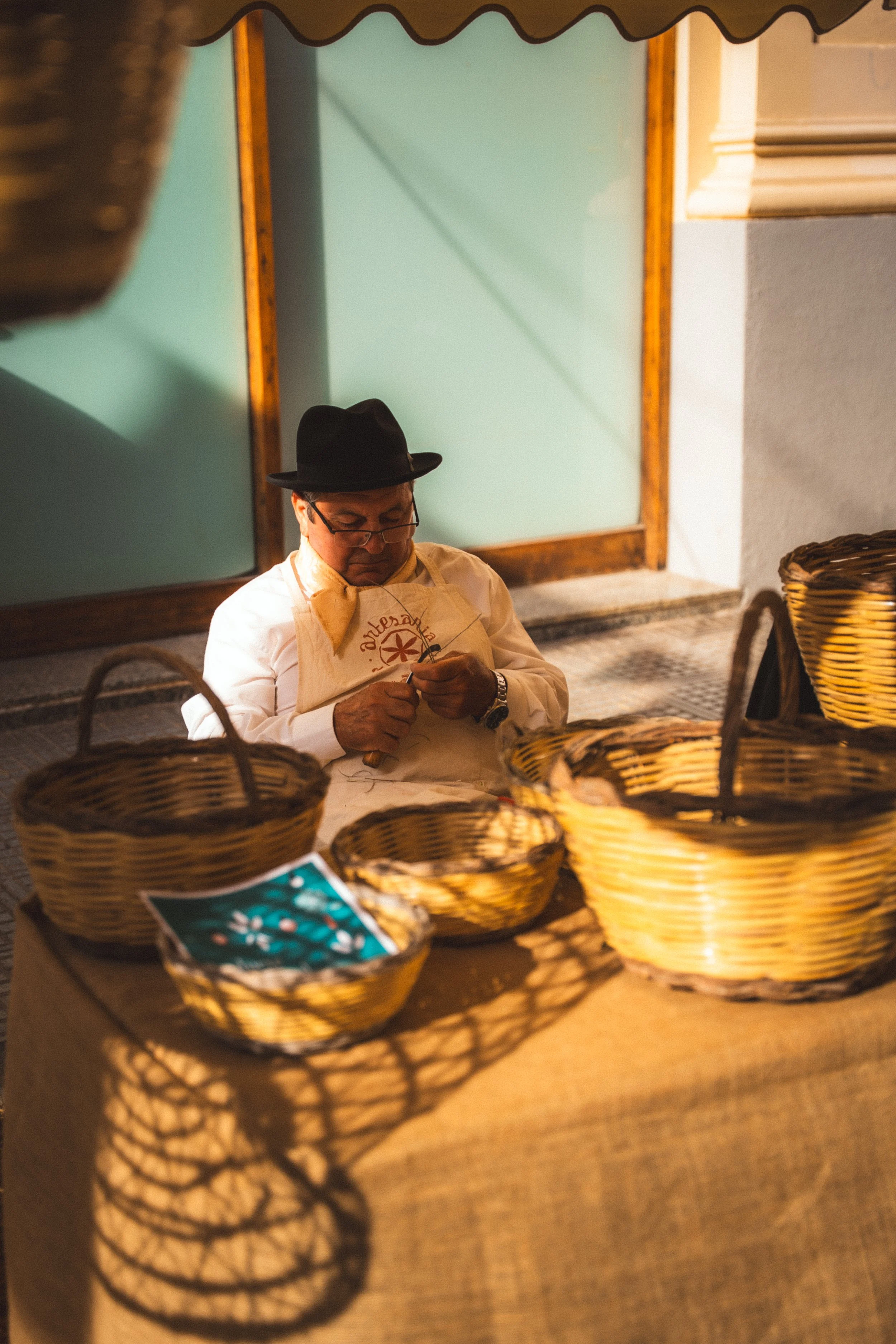 Man in traditionele kleding, met hoed en bril, die borduurt of breit, zittend achter een tafel met manden en tafelkleden, onder een luifel.