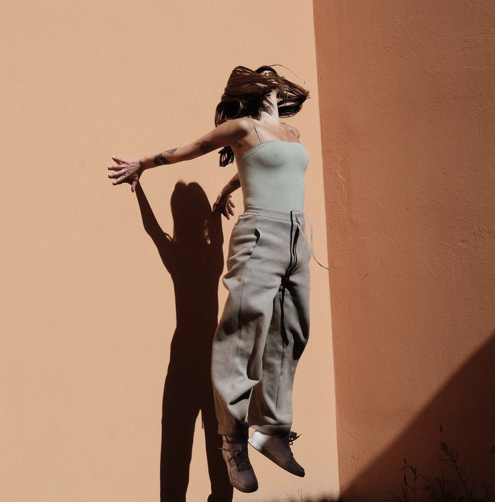 A person with long brown hair, tattoos on their arms, wearing a light grey tank top, baggy beige pants, and sneakers, is captured mid-motion against a two-tone wall with pastel peach and brown colors, casting a shadow on the wall.