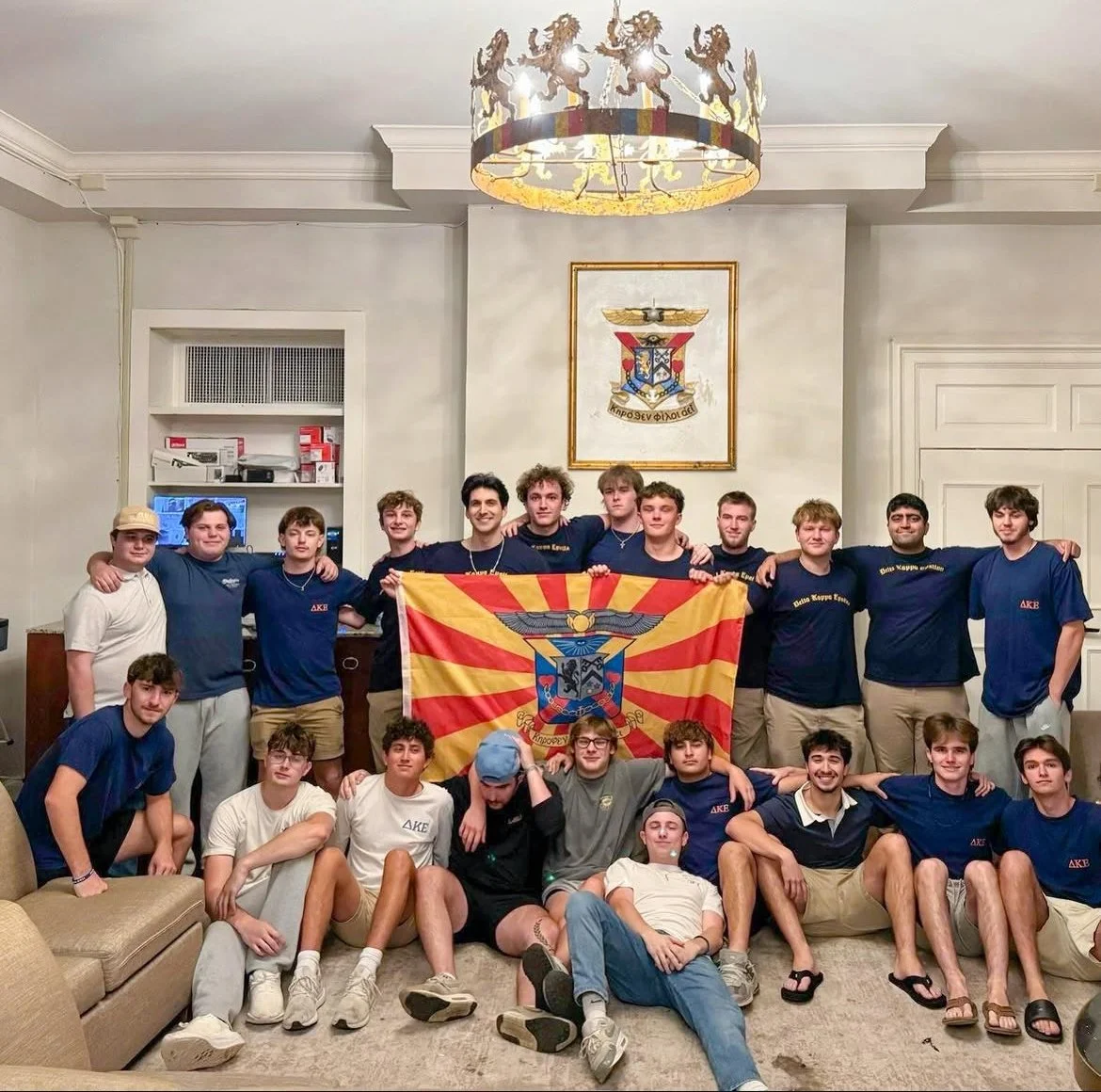 Group of young men in casual clothes posing with a Macedonian flag inside a room with white walls, framed crest, and a decorative ceiling lamp.