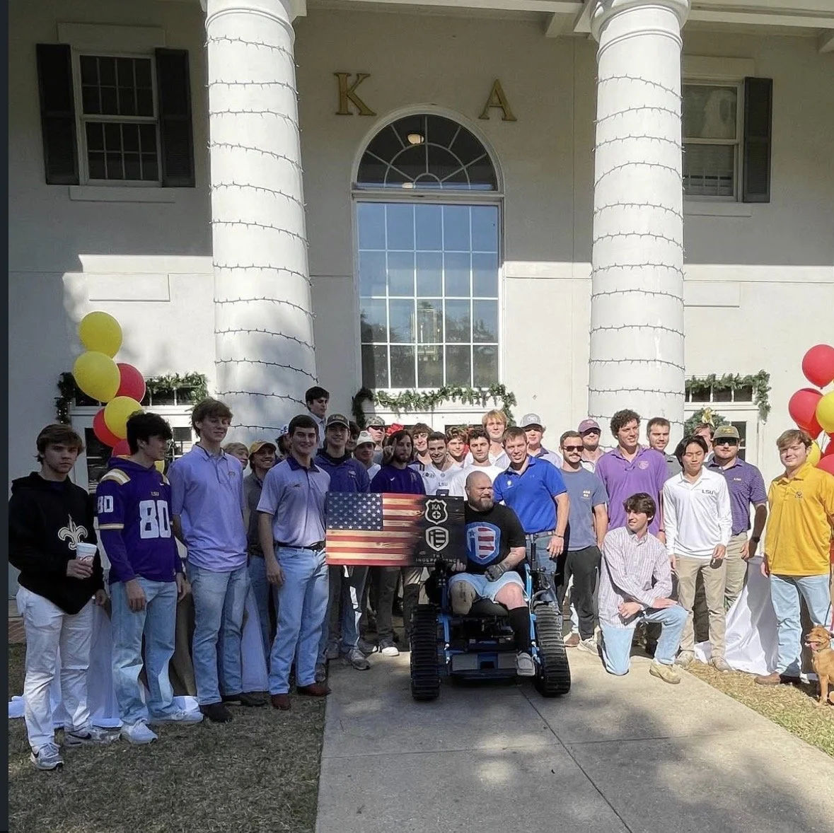 Group of people gathered outside a building with large white columns, holding a flag with an American flag and a logo, with balloons and greenery decoration.