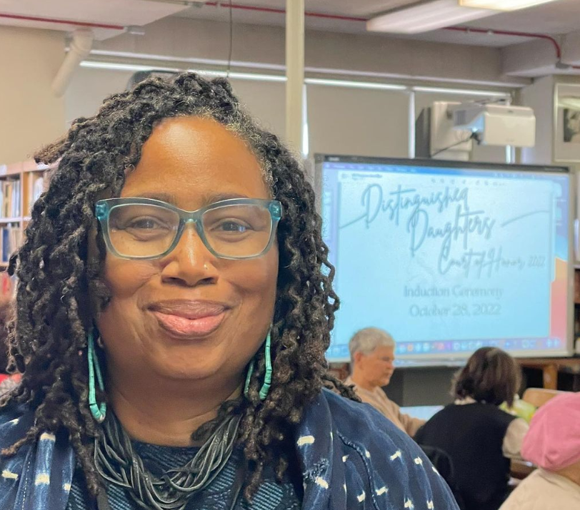 A woman with glasses and earrings smiling in a classroom. Behind her, a presentation screen displays 'Distinguished Daughter of Connecticut' and a date of October 28, 2022, with a few people seated in the background.