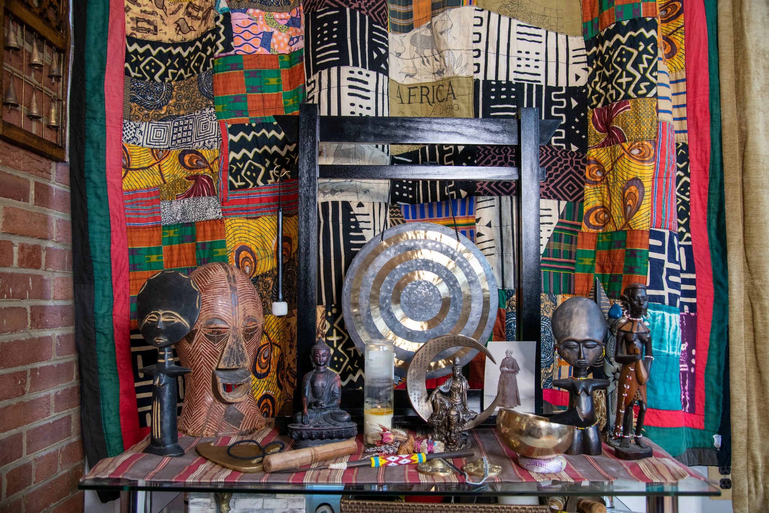 Collection of African artifacts including masks, statues, a gong, and various Buddhist (statues of Buddha and Kuan Yin) spiritual objects displayed on a table with a colorful African textile backdrop.