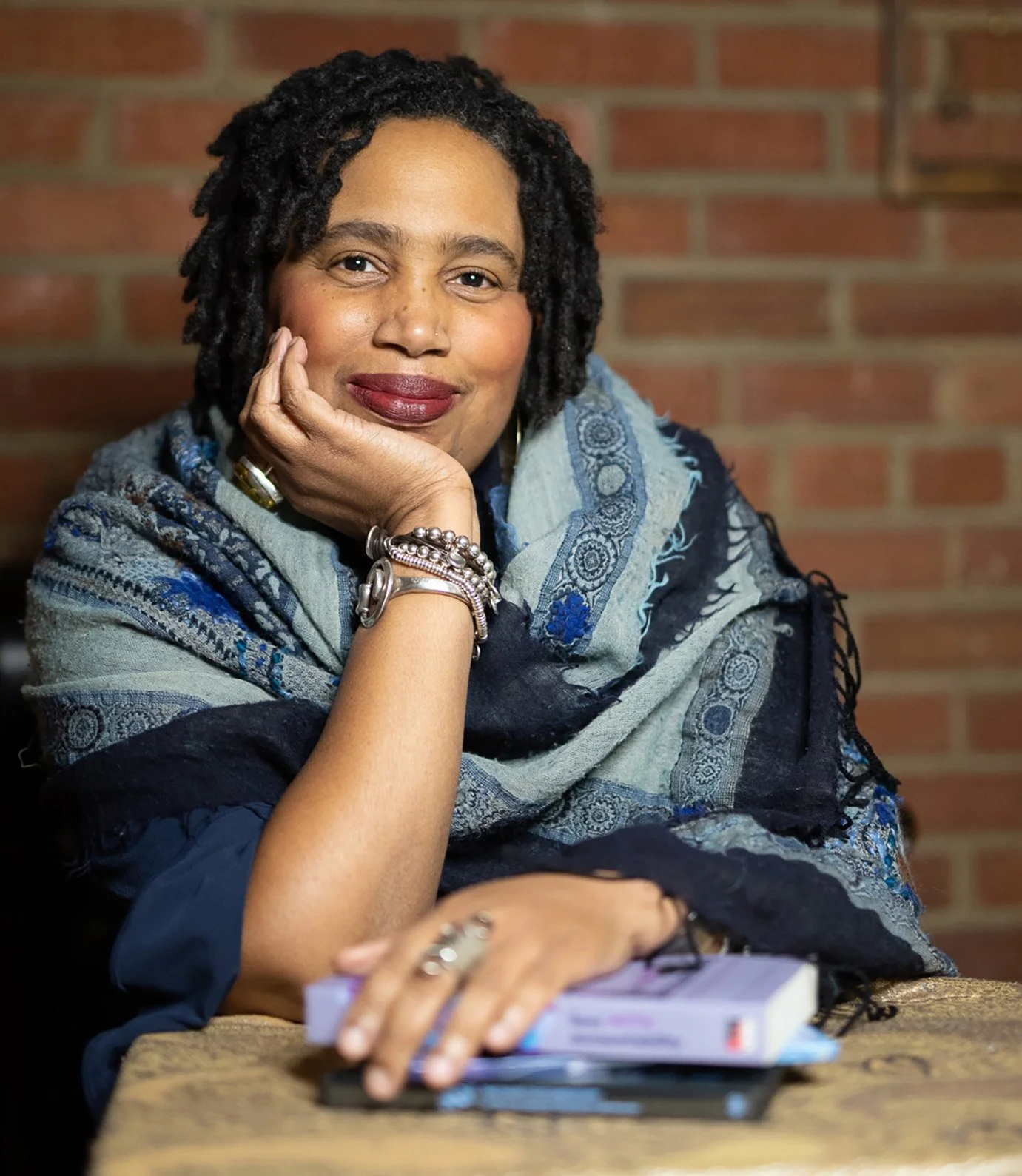 A woman with dark, curly hair and a warm smile, resting her chin on her hand, sitting at a table with a brick wall background, wearing a patterned scarf and jewelry.
