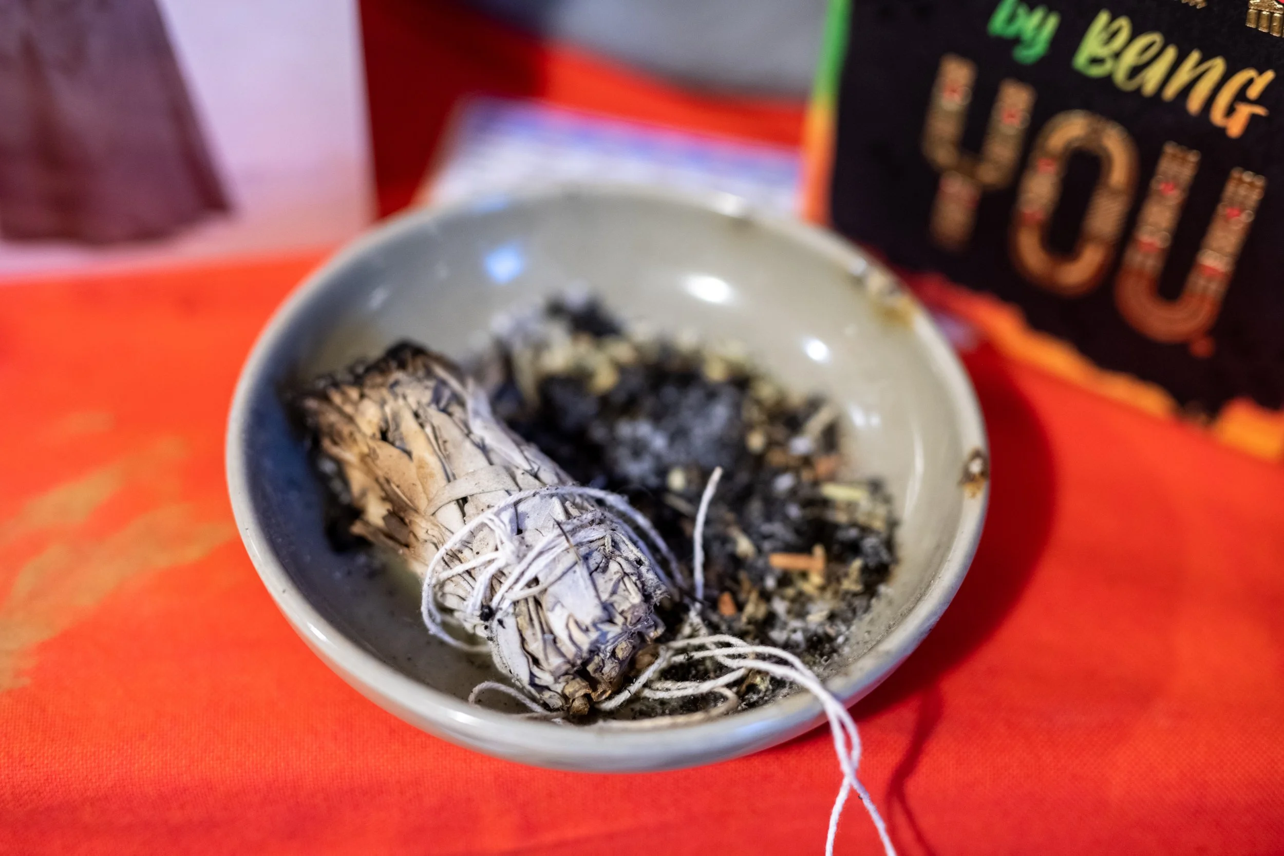 A small bowl containing a bundle of sage and ashes, possibly used for smudging or cleansing rituals, placed on an orange surface with a blurred background.