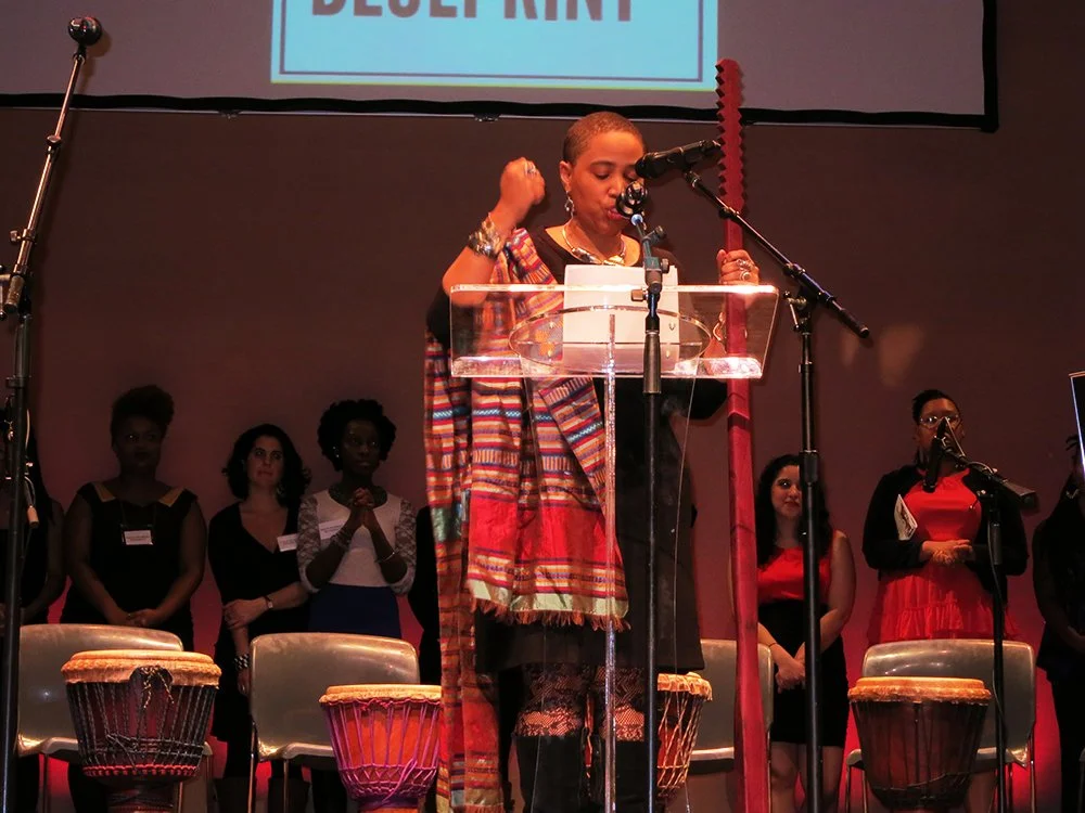 A woman standing at a podium speaking into a microphone on stage, with a group of women standing behind her, some with their hands clasped. There are drums in front of the stage and a large screen above with partially visible text.