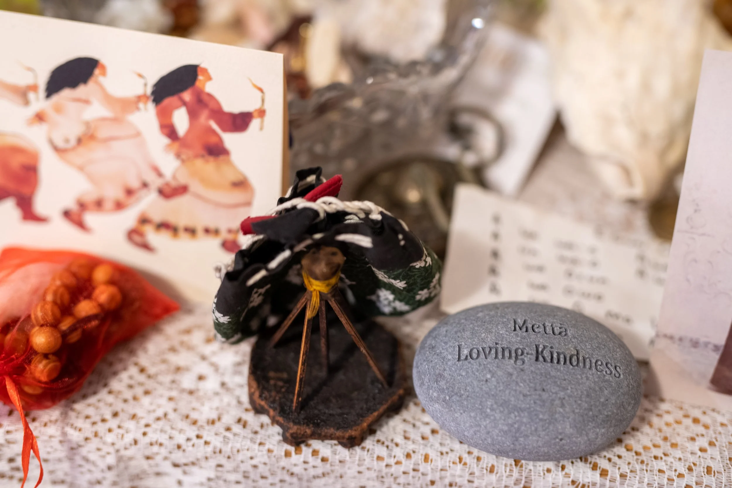 A memorial altar with a gray stone engraved with the words "Metta Loving Kindness," a small black figurine, a picture of traditional dancers, and various notes and items on a lace cloth.