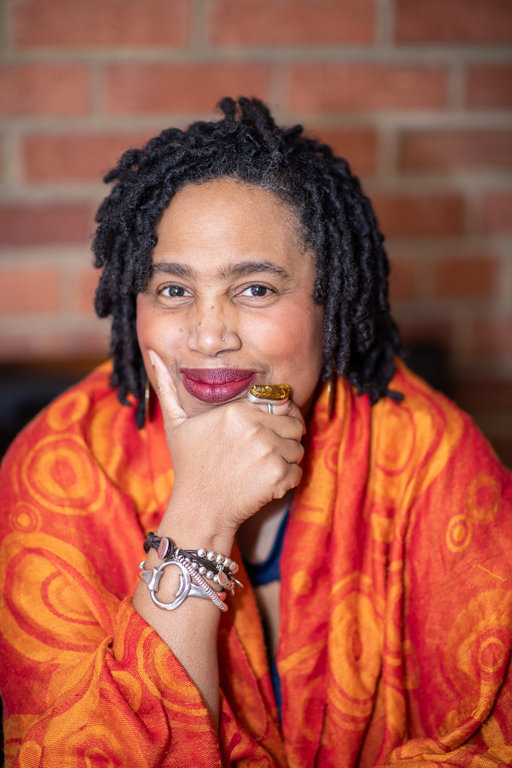 A woman with dreadlocked hair, wearing red lipstick and a hemp nose piercing, sits with her hand resting on her chin, wearing multiple bracelets and a large amber ring, in front of a brick wall.