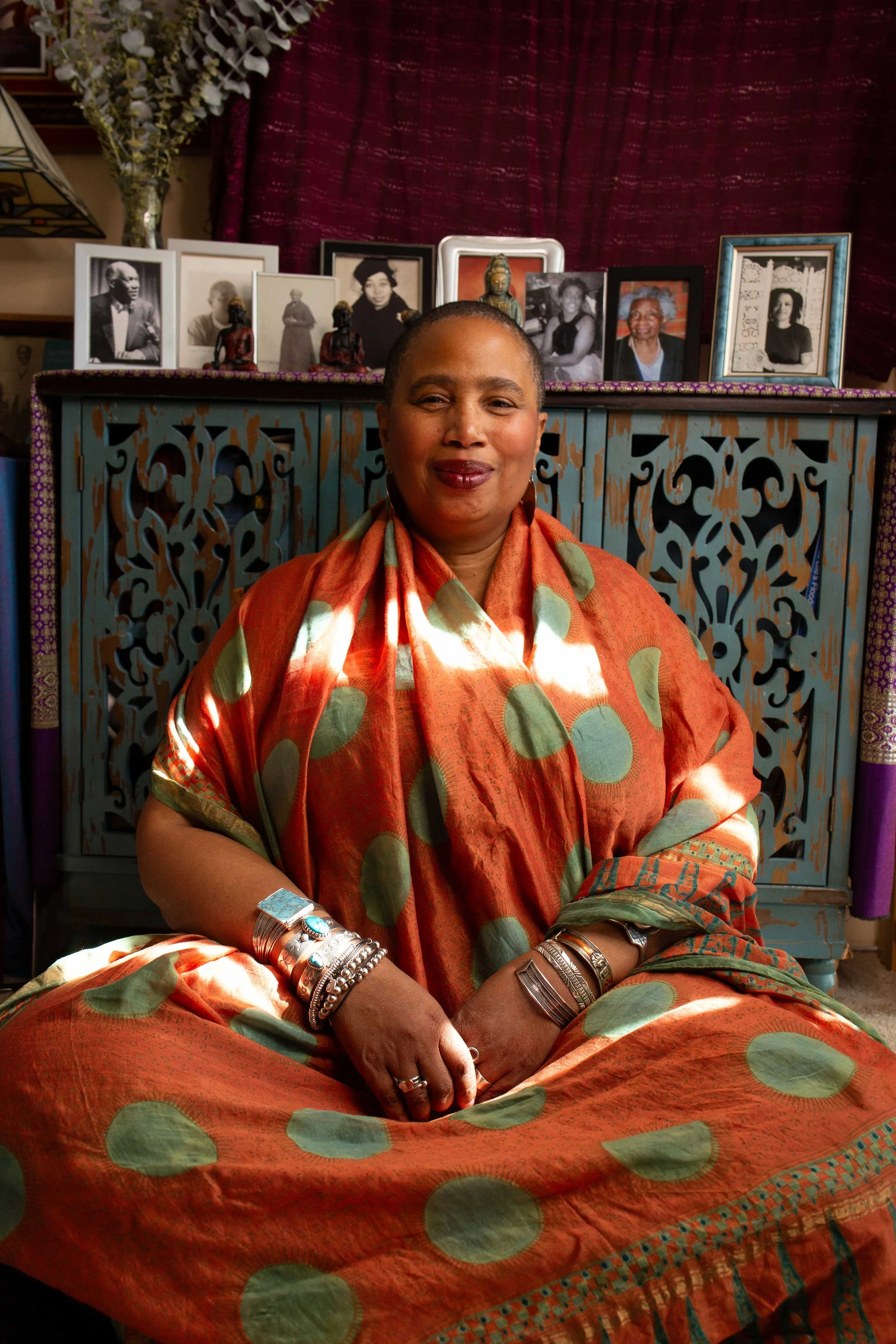 Aishah Shahidah Simmons seated before her ancestral altar, smiling gently, hands folded. Photo by Studio TSHAY
