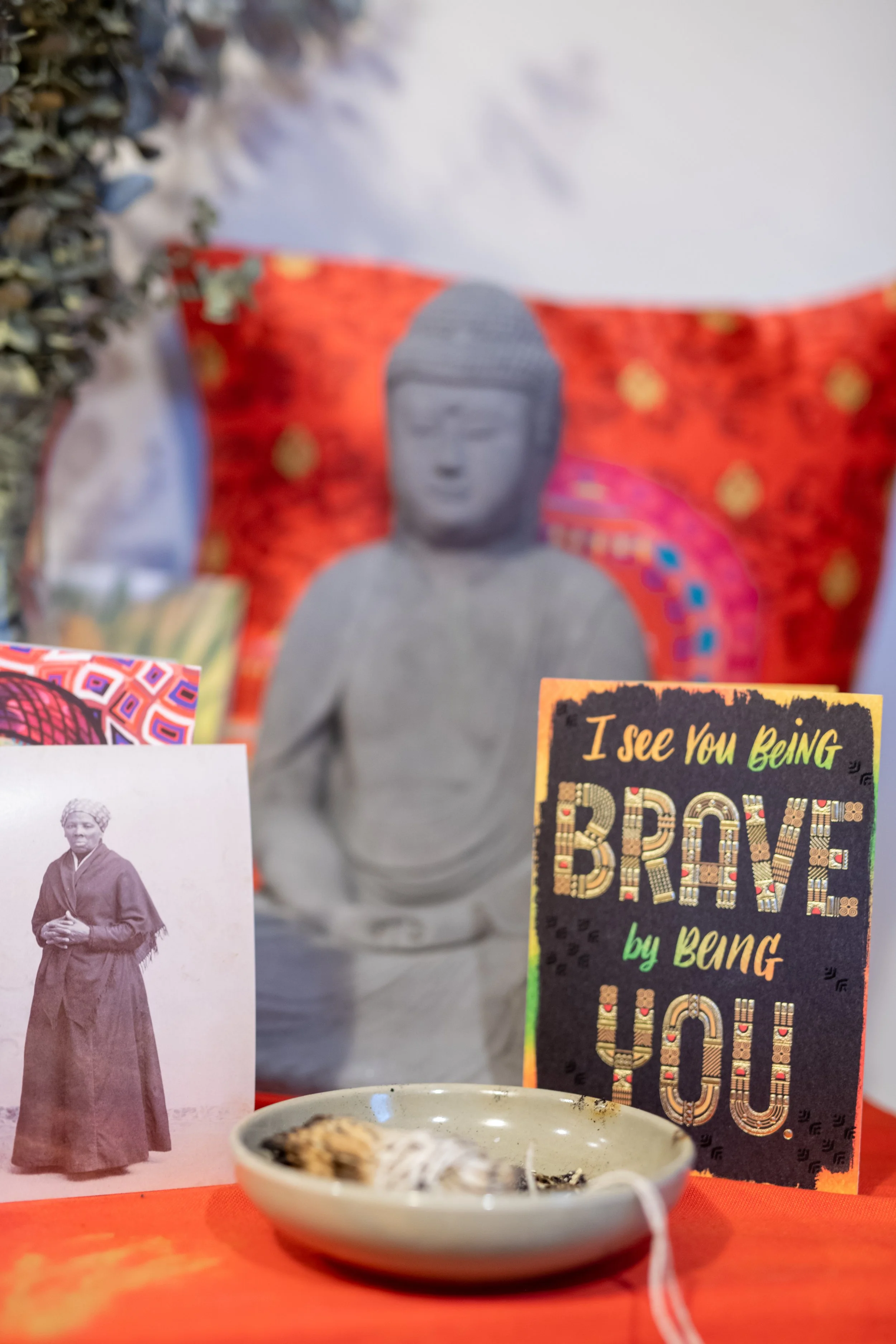 A memorial display with a stone Buddha statue, a black-and-yellow sign that reads 'I see you being brave by being you!', a black and white photo of a woman in traditional attire, a colorful card, and a bowl with some objects, all arranged on a red cloth.