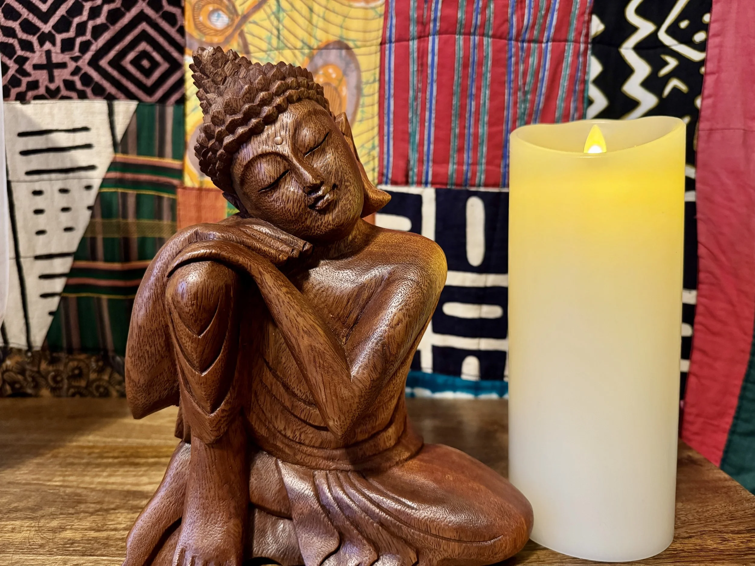 Wooden Buddha statue beside a lit candle, set against a colorful mud cloth backdrop