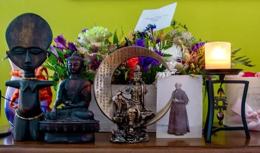 Various statue sculptures and a lit candle on a table with flowers and notes in the background.