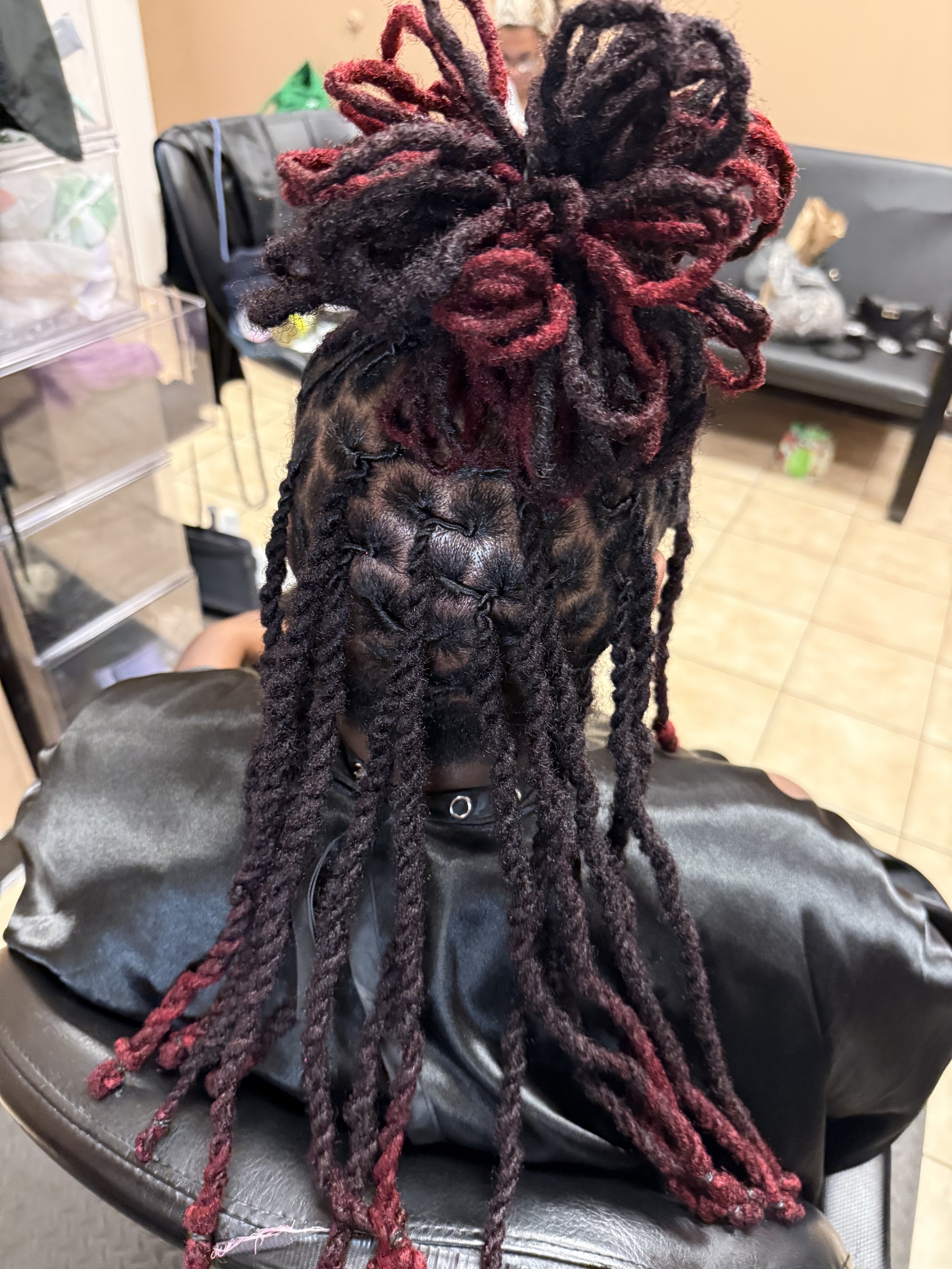 Close-up of a person's head showcasing freshly styled dreadlocks with red and black color. The dreadlocks are thick and well-maintained, arranged in a high updo. The person is seated in a salon or barbershop with a black leather chair and hair stylin