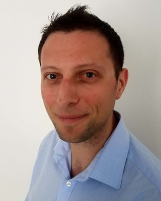 A man with short dark hair and light skin, wearing a light blue collared shirt, smiling slightly in front of a white background.