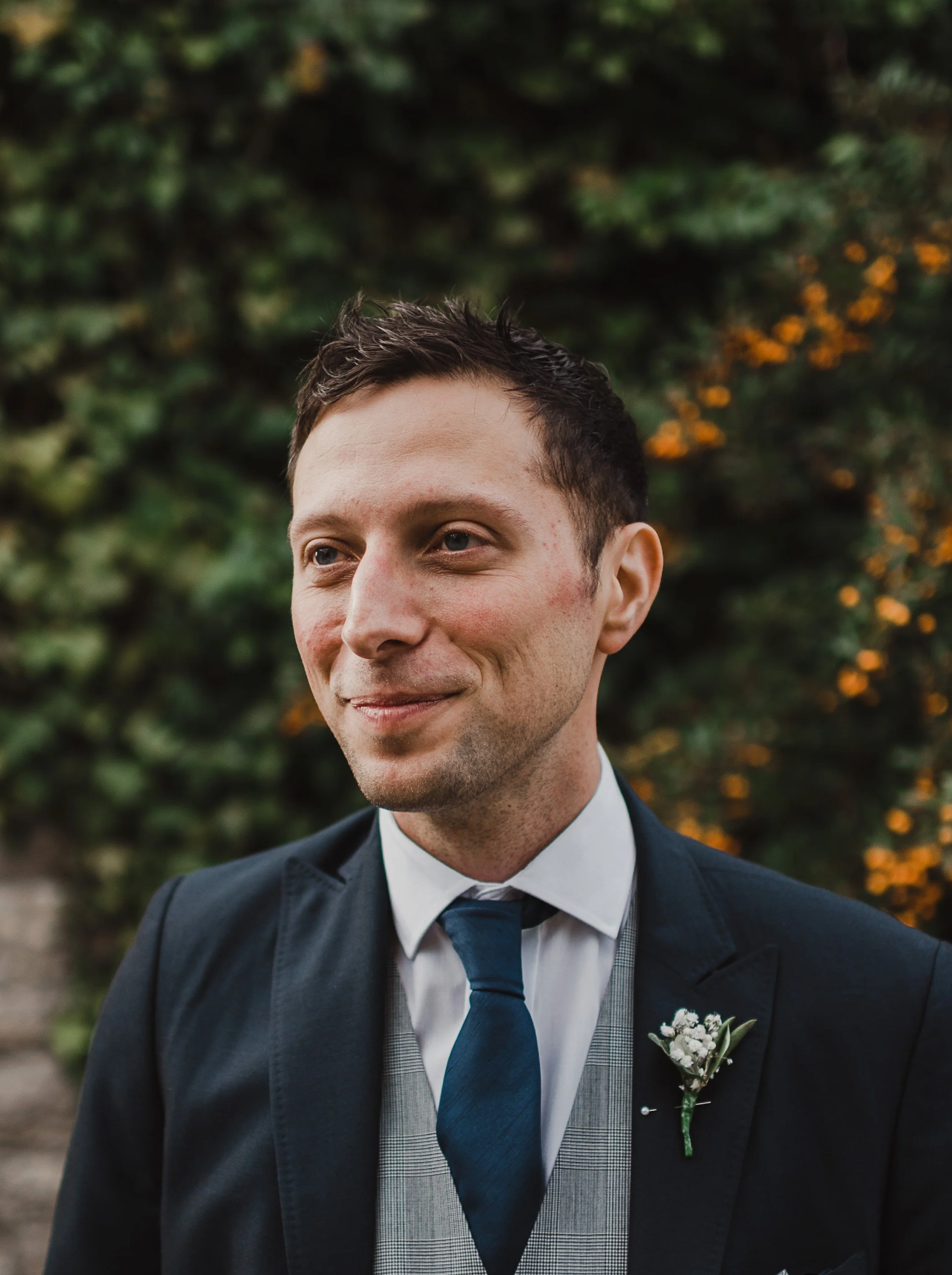 A man in a dark suit with a boutonniere on his lapel, wearing a white shirt and blue tie, smiling outdoors with green foliage and orange flowers in the background.