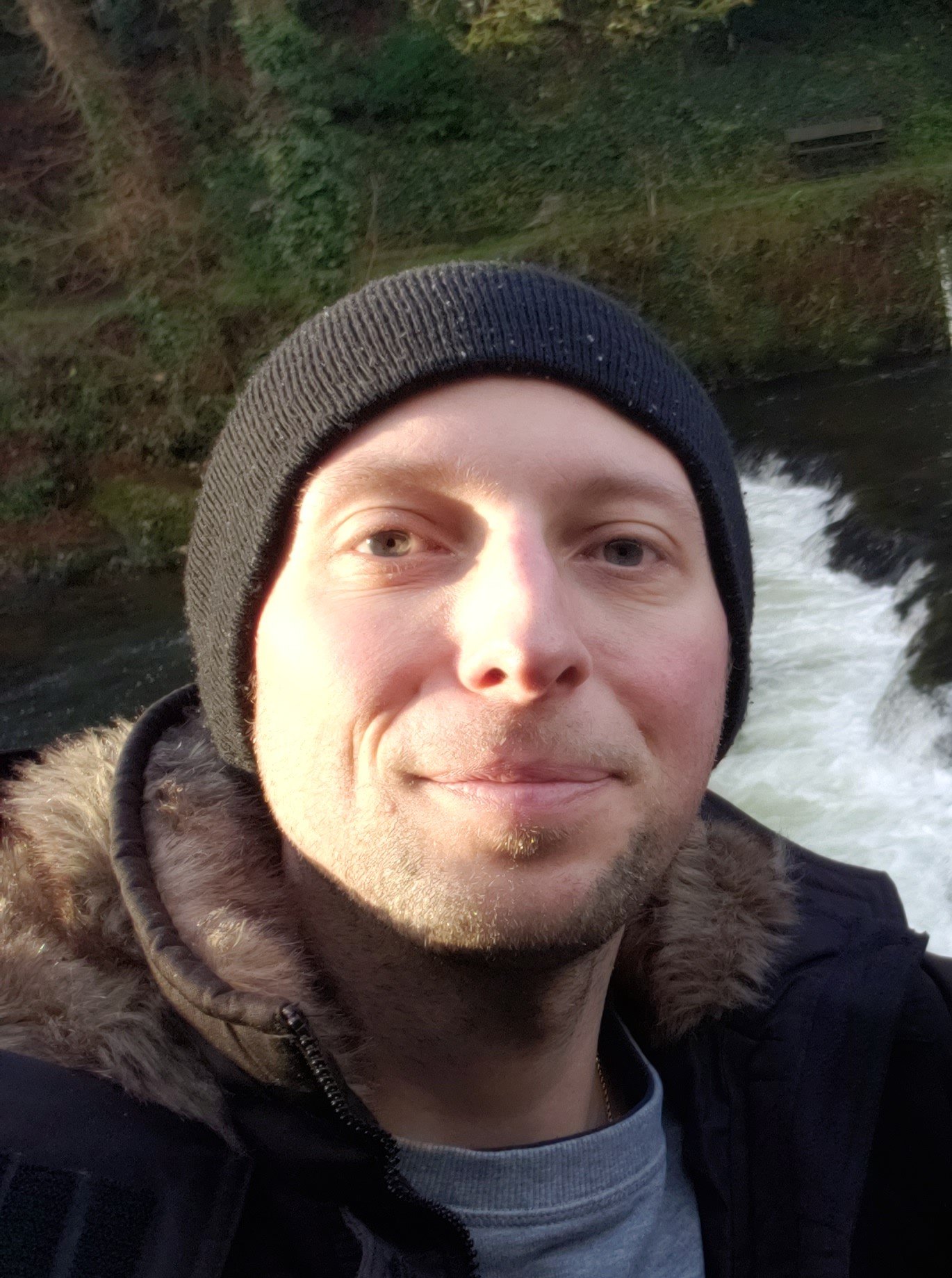 A smiling young man taking a selfie outdoors near a river, wearing a black beanie and a coat with a fur-lined hood.