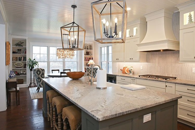 Bright kitchen with white cabinets, large marble island, modern chandeliers, and a dining area with windows, bookshelves, and cozy chairs.
