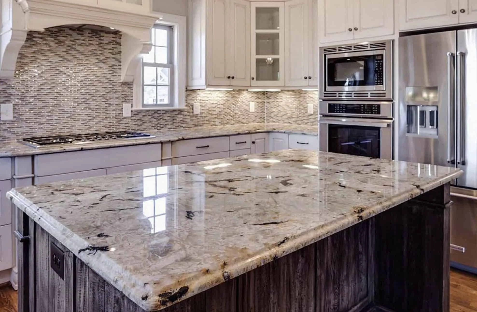 Modern kitchen with granite island, white cabinets, tiled backsplash, stainless steel appliances, and a window.