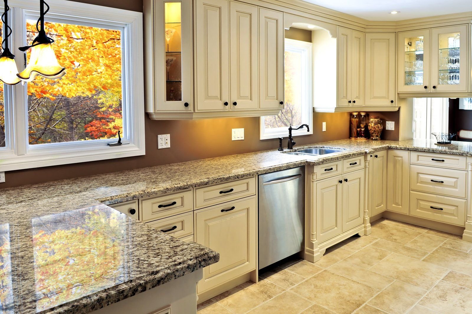 Kitchen with cream-colored cabinets, granite countertops, a stainless steel dishwasher, and windows showing fall foliage outside.