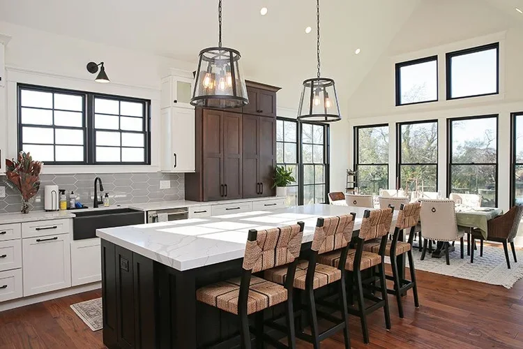 Modern kitchen with large island, white and dark wood cabinetry, pendant lighting, and dining area with chairs near large windows.