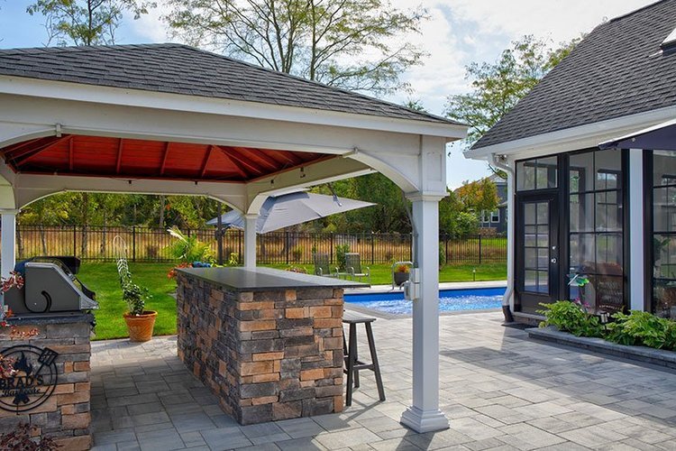 Outdoor patio area with a stone bar, grill, and seating, covered by a pavilion with a red ceiling, next to a pool and a sunroom with large windows, in a backyard with a wooden fence and trees.