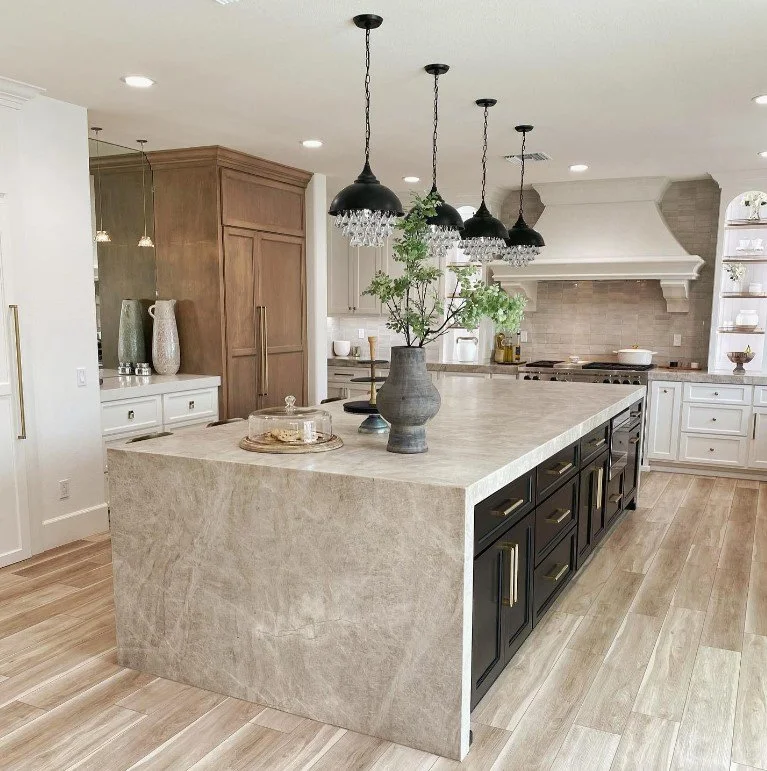 Modern kitchen with a large beige marble island, black cabinets, and white cabinets in the background. Pendant lights hang above the island, which holds a vase with greenery and a glass cake stand.