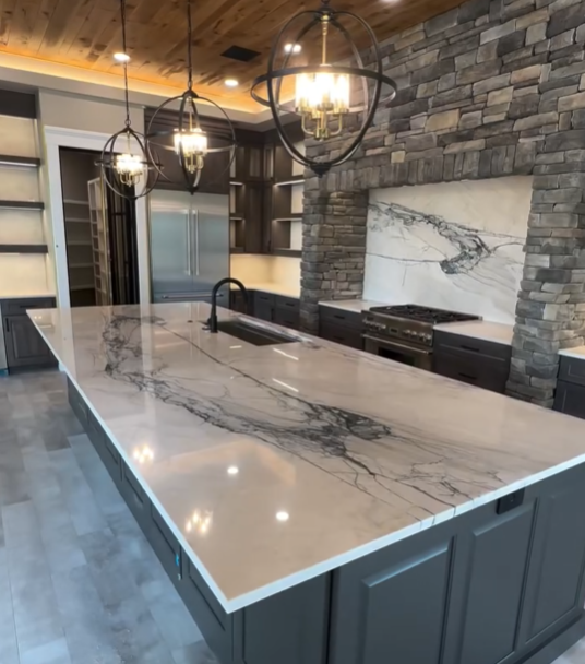 Modern kitchen with a large marble island, black cabinetry, stone accent walls, and pendant lighting.