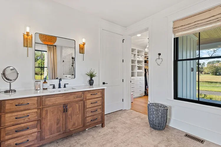 A bathroom with a wooden vanity, mirror, and wall-mounted lights, a window with a view of greenery, and an open door leading to a walk-in closet.