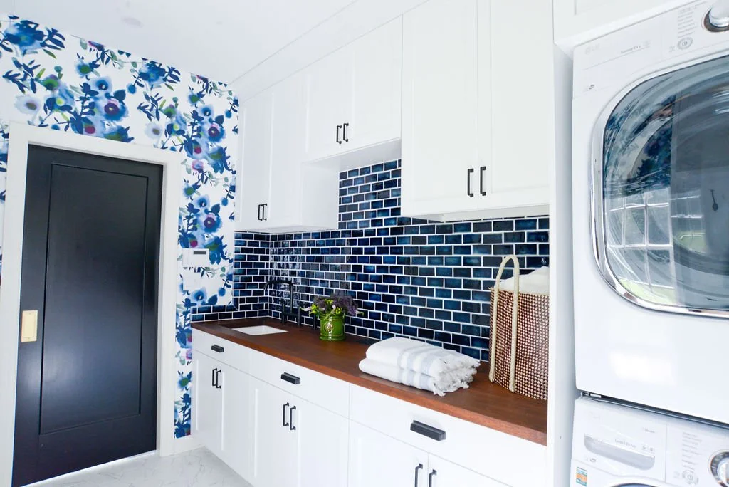 Laundry room with white cabinets, black door, dark blue subway tile backsplash, wooden countertop, and stacked washer and dryer. Decor includes a green plant, white towels, and a basket. Floral wallpaper on adjacent wall.