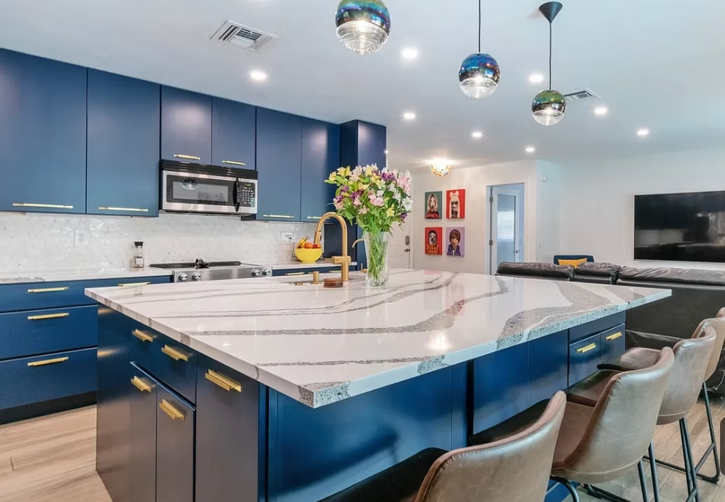 Modern kitchen with blue cabinets and gold hardware, large marble island with a vase of flowers, pendant lights, and a living room area with a TV and artwork in the background.