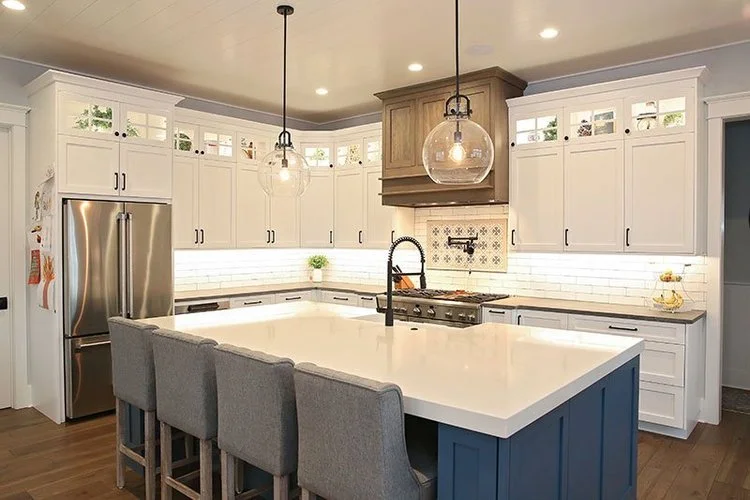 Modern kitchen with white cabinets, stainless steel refrigerator, central island with blue base, gray chairs, pendant lights, and a backsplash with decorative tiles.