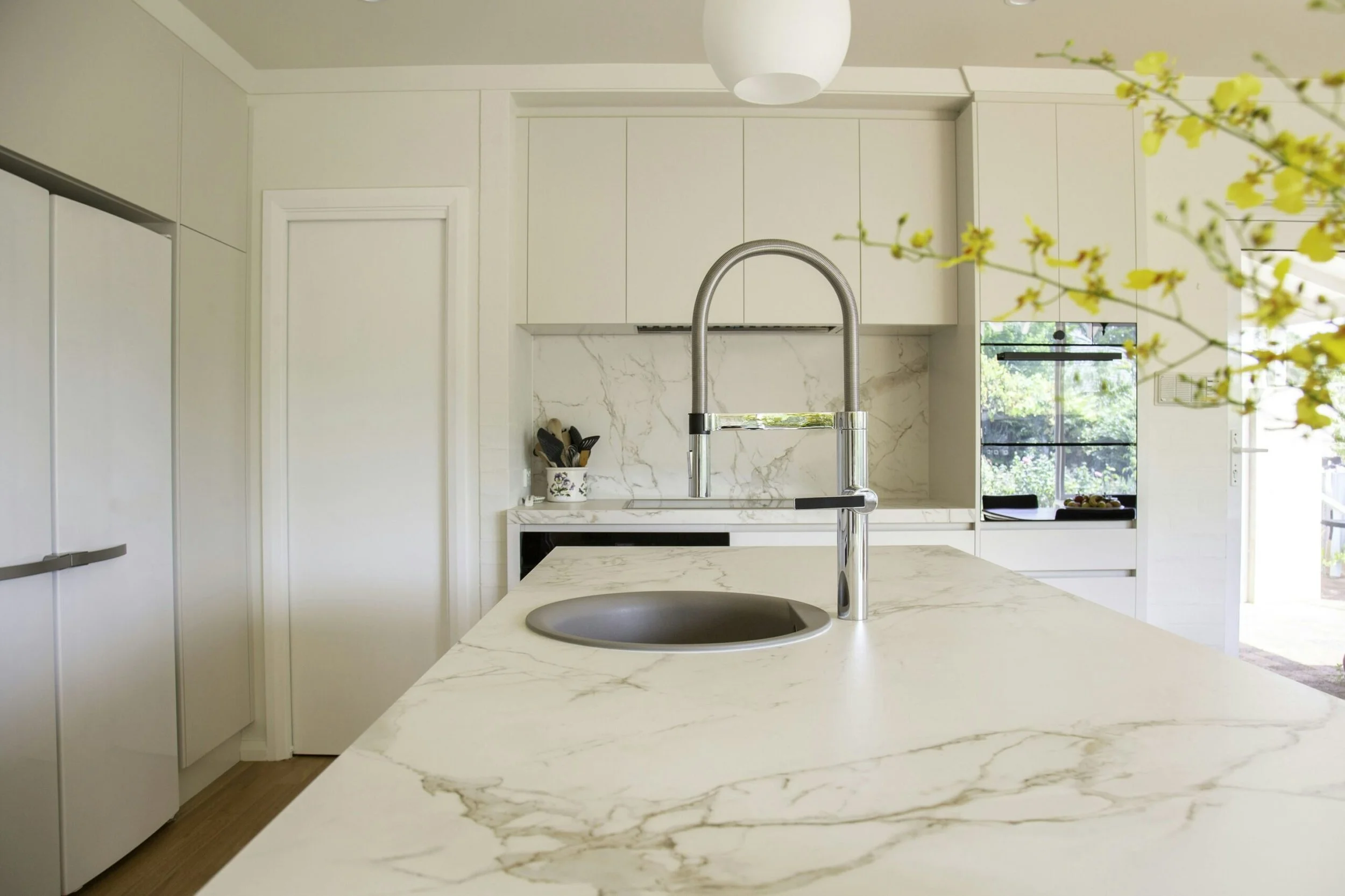Modern kitchen with white marble countertops, a built-in sink, a shiny silver faucet, white cabinets, a window with greenery outside, and a branch with yellow flowers in the foreground.