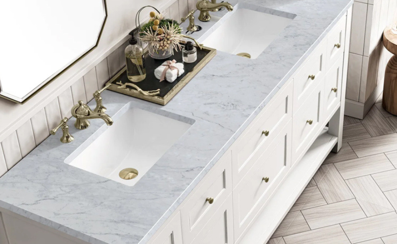 A white bathroom vanity with a marble countertop, two undermount sinks, and gold faucet fixtures. There are small jars and decorative items on a tray near the left sink, a mirror above the vanity, and light-colored wooden flooring.