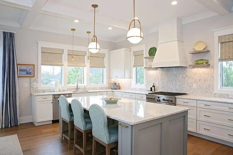 Modern kitchen with white cabinets, marble countertops, and pendant lighting. Four blue upholstered chairs around a marble island, with windows and decorative shelves.