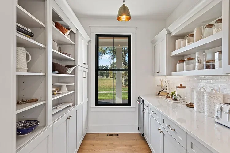 A bright, white kitchen with open shelves and cabinets, a large black-framed window showing a green outdoor scene, and a wooden floor. Gold pendant light hanging from the ceiling.