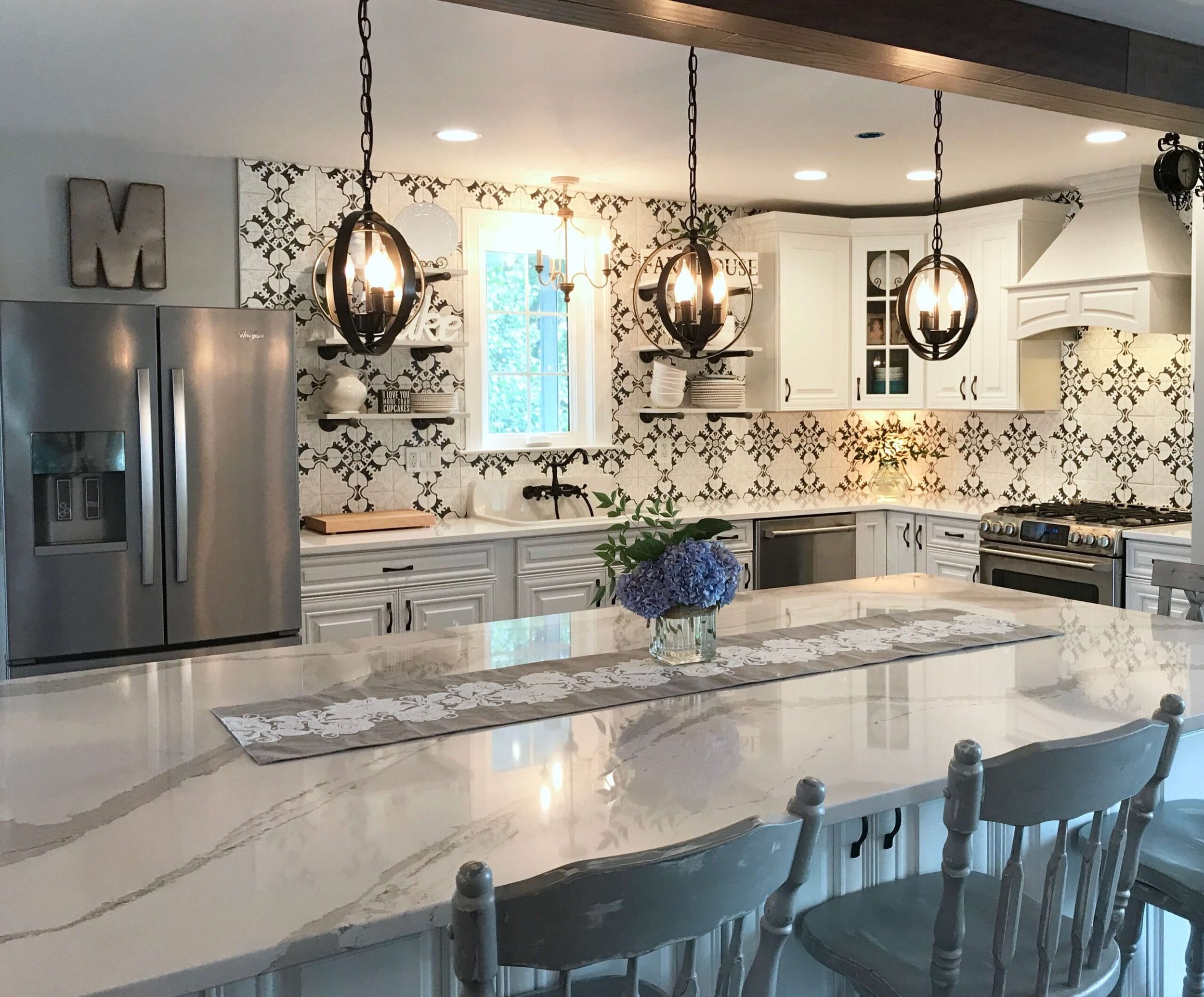 A bright kitchen with white cabinets, black and white patterned tile backsplash, stainless steel refrigerator, and a large central island with a white marble countertop. Hanging pendant lights and a window above the sink add to the space's charm. A v