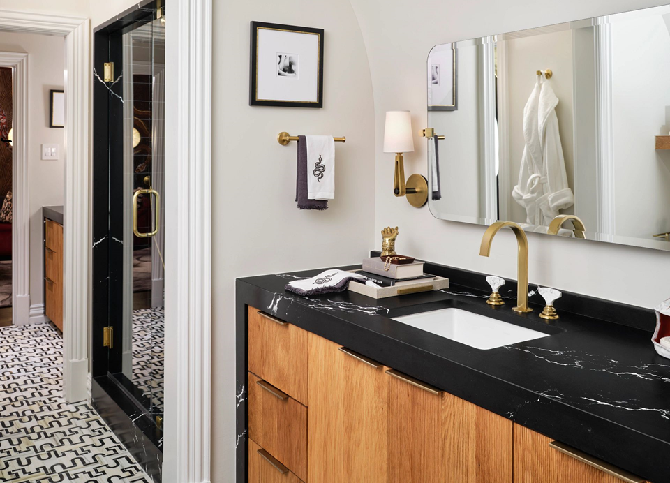 Modern bathroom with a black marble countertop featuring gold fixtures, a wooden vanity, and a large mirror. There are framed pictures, a towel rack with a white towel, and a shower with a black door in view.