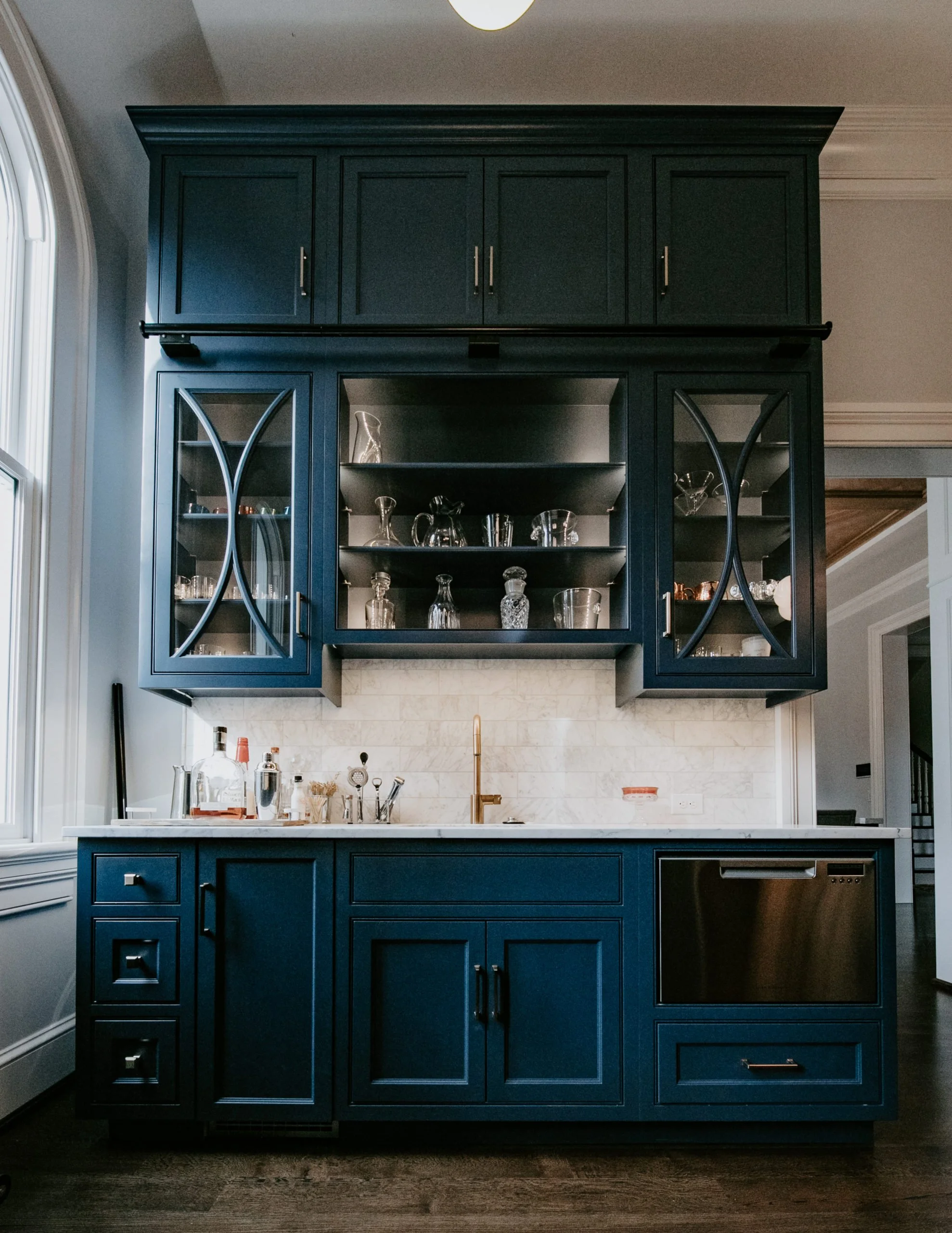 Dark blue kitchen cabinet with glass doors displaying glassware behind a white marble countertop with a gold faucet, next to a black dishwasher.