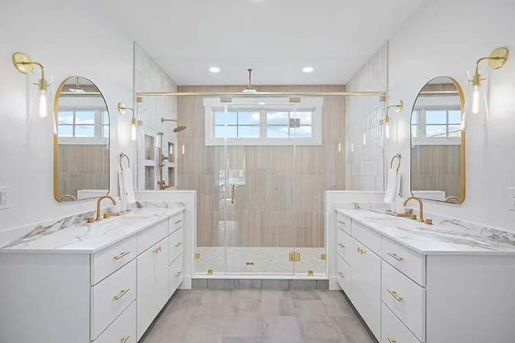 Modern bathroom with twin white vanities featuring marble countertops and gold fixtures, large framed mirrors, and a walk-in shower with clear glass door and gold hardware. Light-colored walls and flooring with a window and ceiling lights.