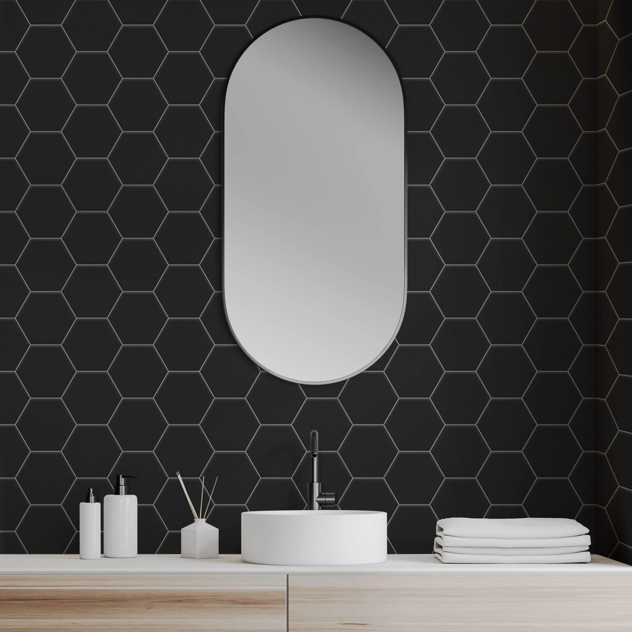 Modern bathroom vanity with a white oval sink, black hexagonal tiled wall, oval mirror, white towels, soap, and reed diffuser on a light wood cabinet.