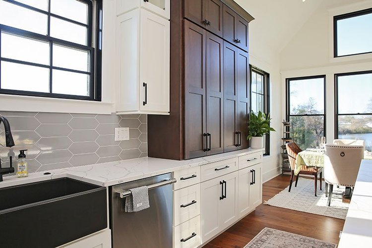 Modern kitchen with white and dark wood cabinets, gray hexagonal tile backsplash, black sink, and large windows overlooking an outdoor patio and water view.