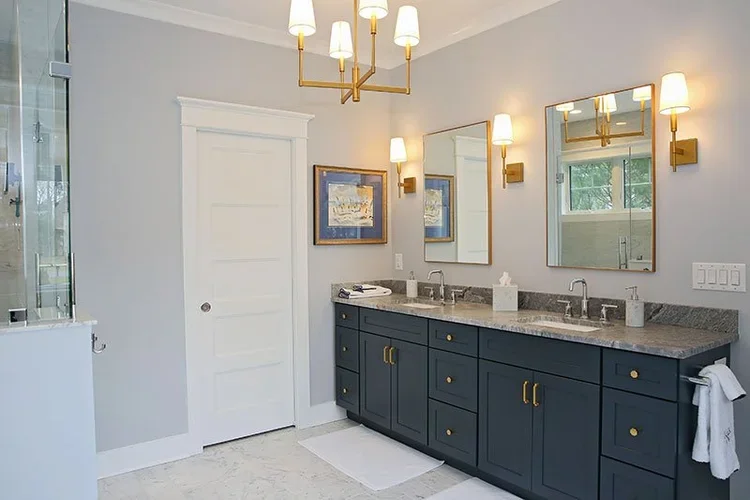 A modern bathroom with double sinks, navy blue vanity, marble countertops, two mirrors, wall-mounted lights, and a large window.