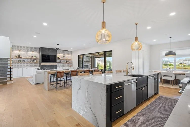 Open-concept modern kitchen and living room with white cabinets, black accents, pendant lighting, and a dining area near large windows.