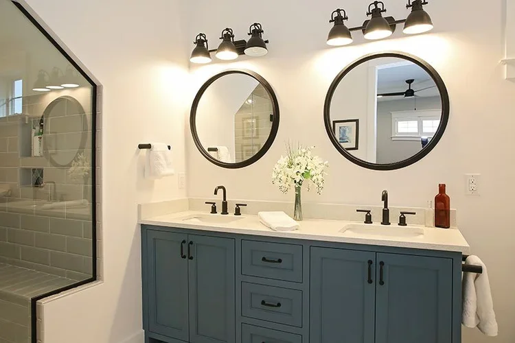 Bathroom with a blue vanity, two round mirrors, black fixtures, and a white countertop with a vase of white flowers and decorative bottle, bright lighting