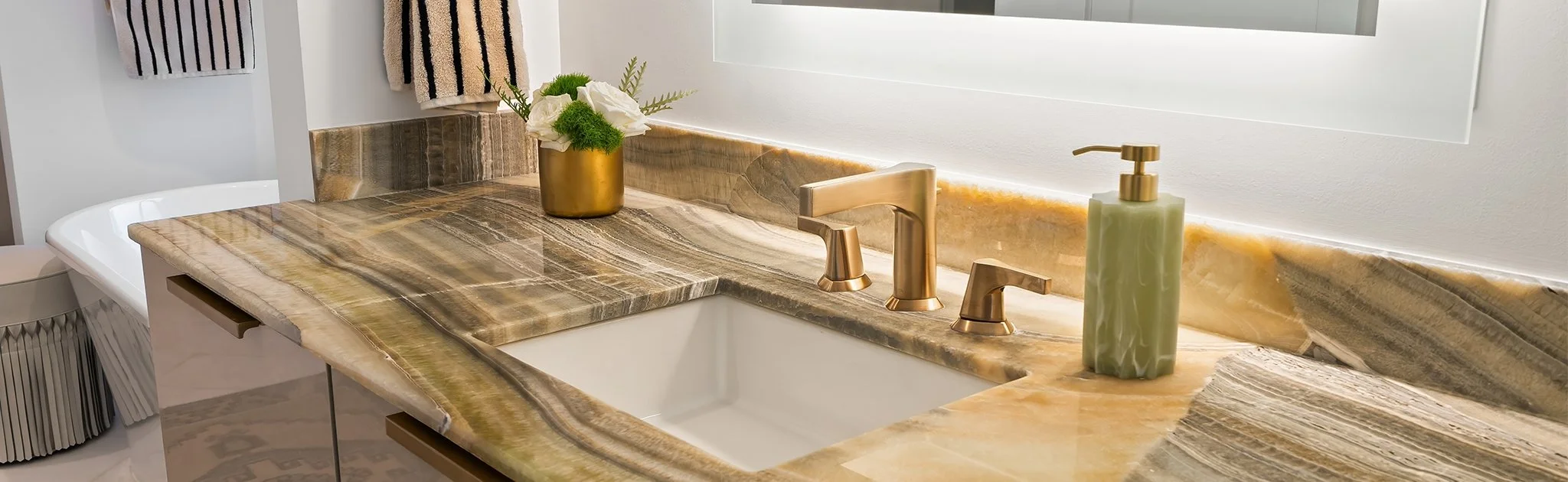 Close-up of a bathroom vanity with a marble countertop, gold faucet, a gold vase with white flowers, and a green soap dispenser.