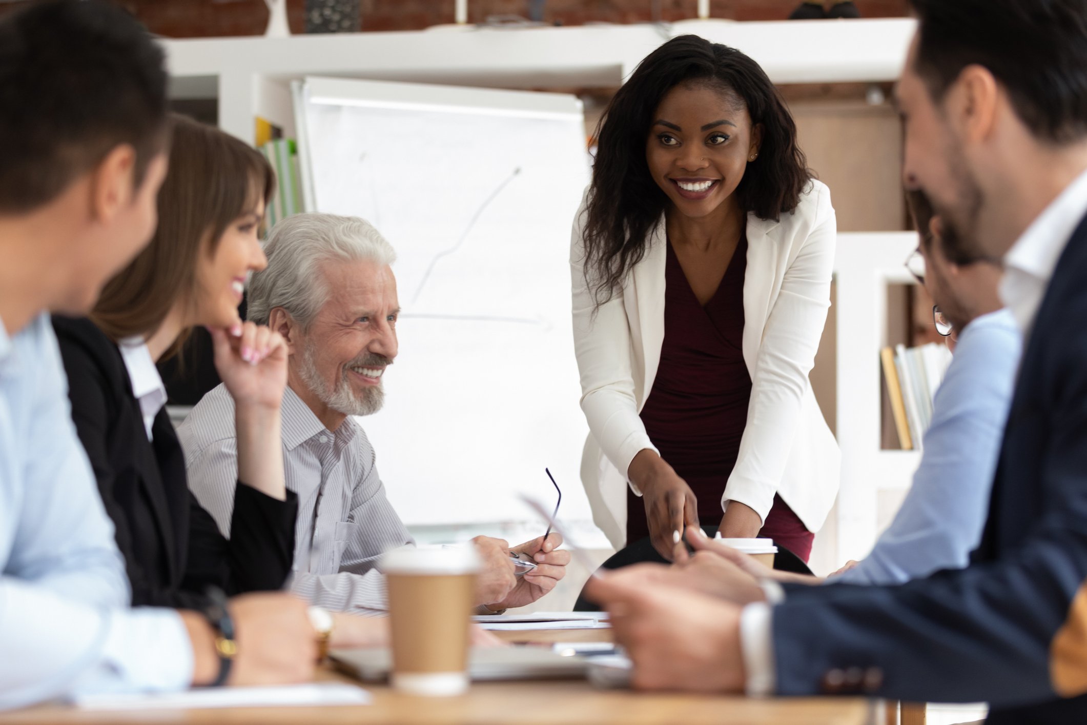 Business team meeting with diverse colleagues, including African American woman presenting and smiling, and older man listening, in a modern office conference room.