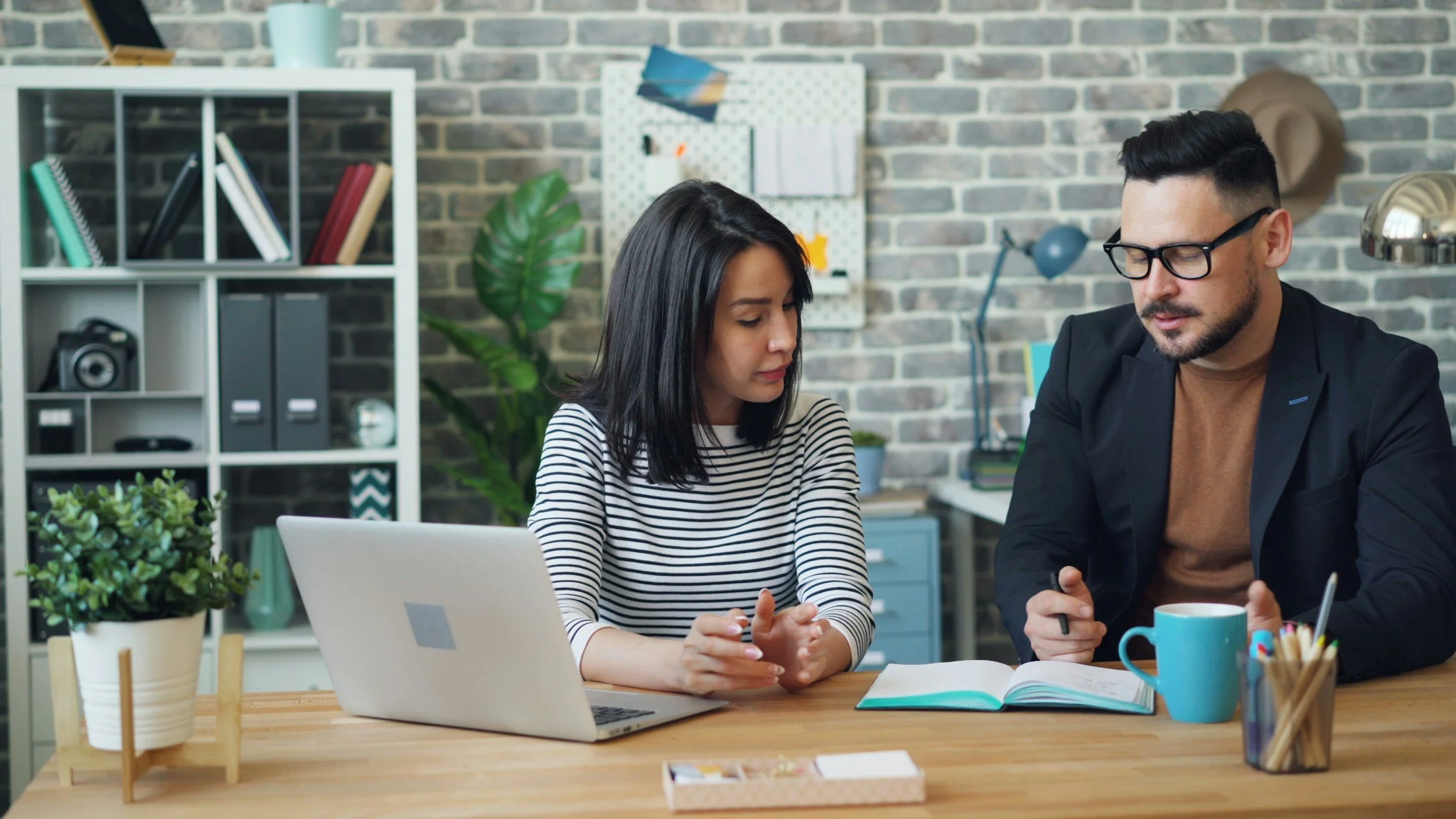 Two people sitting at a wooden table in an office, looking at a phone together. The woman has dark hair and is wearing a striped shirt. The man has short dark hair, glasses, and is wearing a black blazer. There are books, a laptop, a mug, and office supplies on the table. Behind them is a background with a brick wall, a bookshelf, a white pegboard with stationery, plants, and a hat on the wall.
