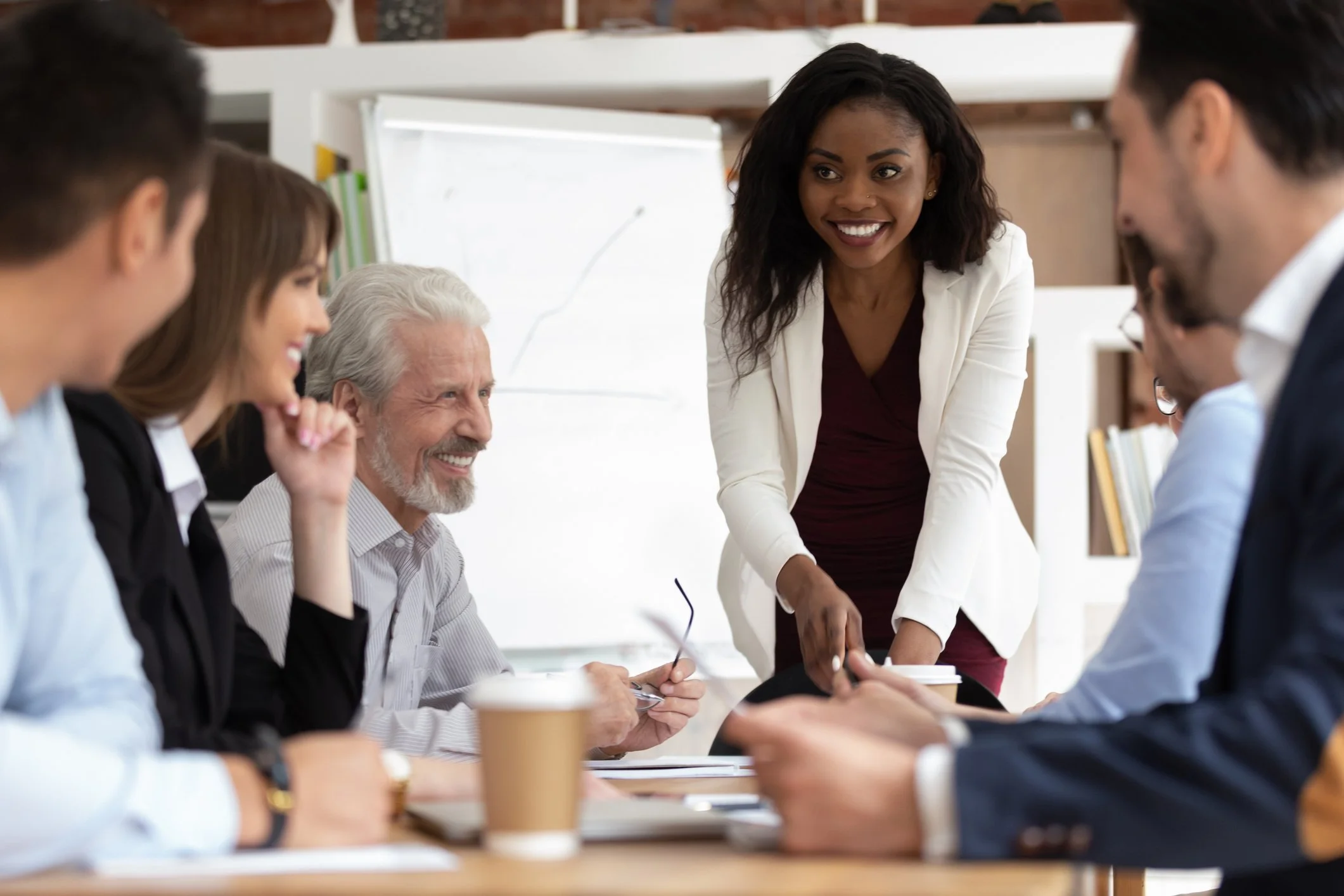 A group of diverse people in a meeting room, with a woman standing and smiling while talking to others seated at the table.