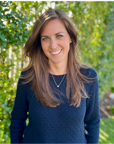 A smiling woman with long brown hair, wearing a navy blue sweater and a necklace, standing outdoors with green trees in the background.