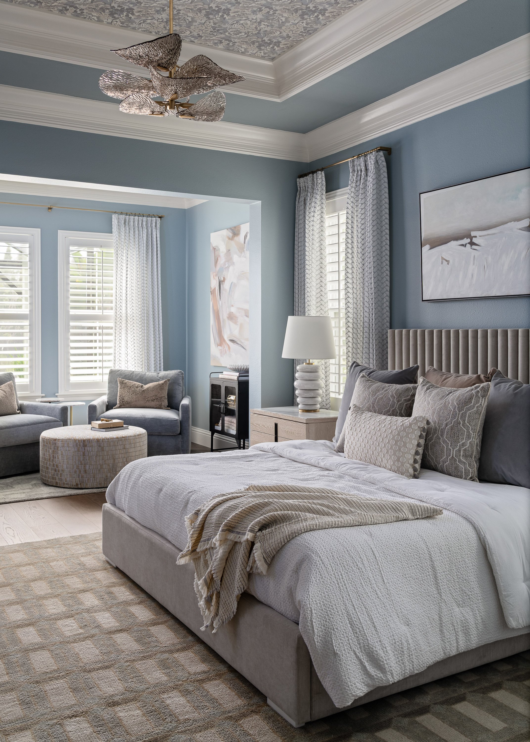 A modern bedroom with light blue walls, white crown molding, and a ceiling chandelier with leaf-like metallic design. There is a large bed with a tufted headboard and neutral bedding, a white nightstand with a decorative lamp, and a cozy sitting area with two armchairs, a small ottoman, and abstract art on the walls. Multiple windows with white blinds and patterned curtains let in natural light.